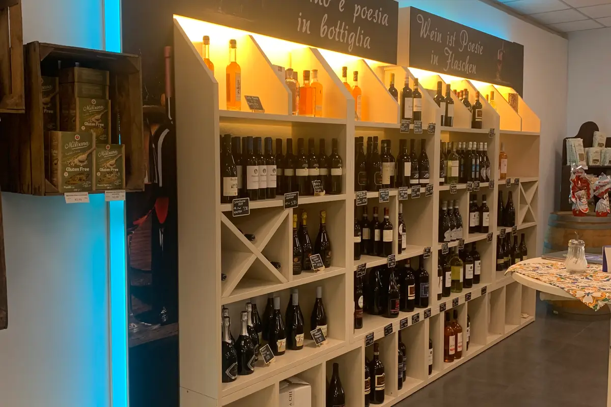 Raft cu diverse sticle de vin și băuturi spirtoase, deasupra a două pancarte cu textele "Wine is poetry in bottles" și "Wine is poetry in bottiglia".