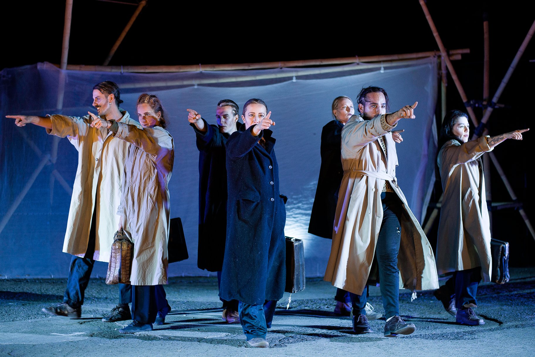 Scena este albastru închis. Șase persoane în paltoane lungi, deschise la culoare, stau la rând și arată cu degetele în direcții diferite. Unii țin pungi în mâini.