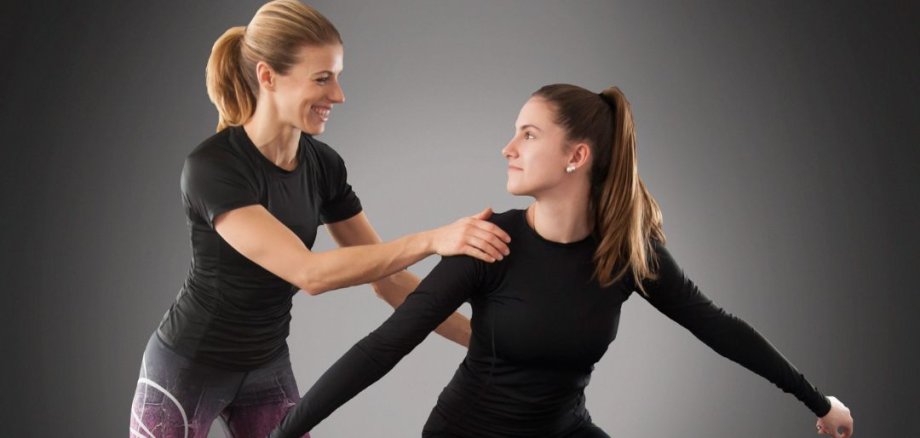 vhs: Brain Gym® pentru adulți Două femei îmbrăcate în ținute sportive; una stă într-o poziție de genuflexiune largă, cu brațele întinse, iar cealaltă îi atinge umărul pentru a o susține