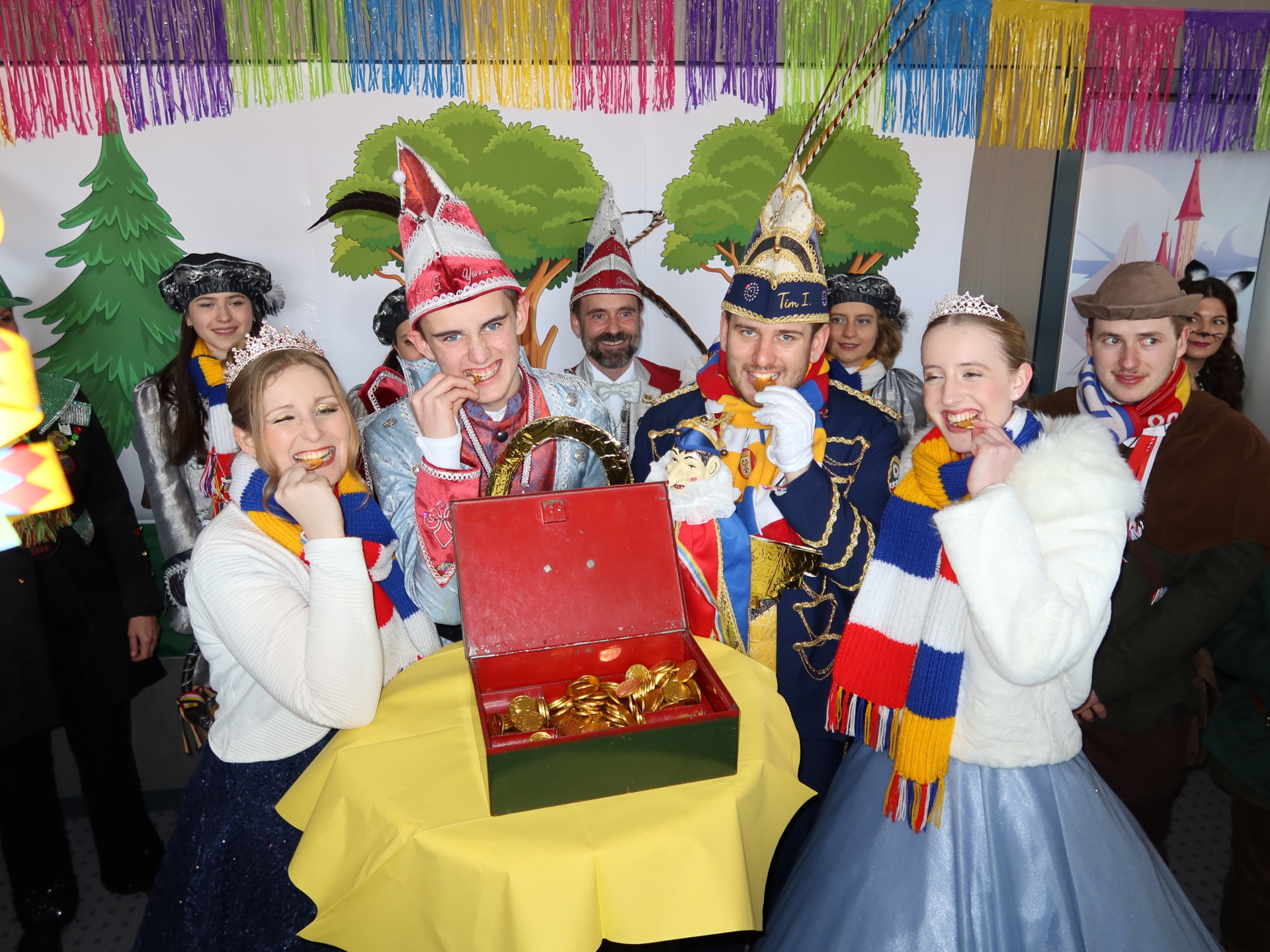 Șase persoane în ținute festive, inclusiv un prinț și o prințesă, stau în jurul unei mese rotunde cu o cutie verde. În fundal se pot vedea decorațiuni colorate și siluete de pom de Crăciun.