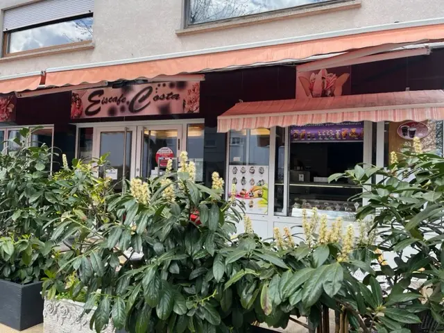 Кафе-мороженое с надписью "Eiscafe Costa" за зелеными растениями в вазонах