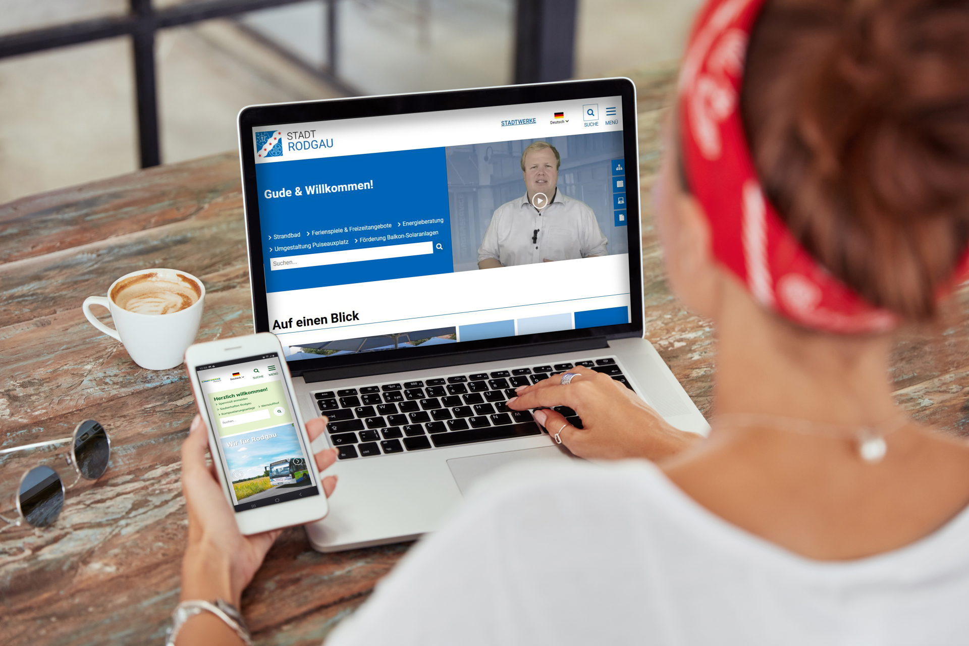 Bir kişi ahşap bir masada oturmuş dizüstü bilgisayar ve akıllı telefon kullanıyor. Dizüstü bilgisayar ekranında Rodgau kasabasının web sitesi ve "Gude & Willkommen!" yazısı görülmektedir. Dizüstü bilgisayarın yanında bir fincan kahve var.