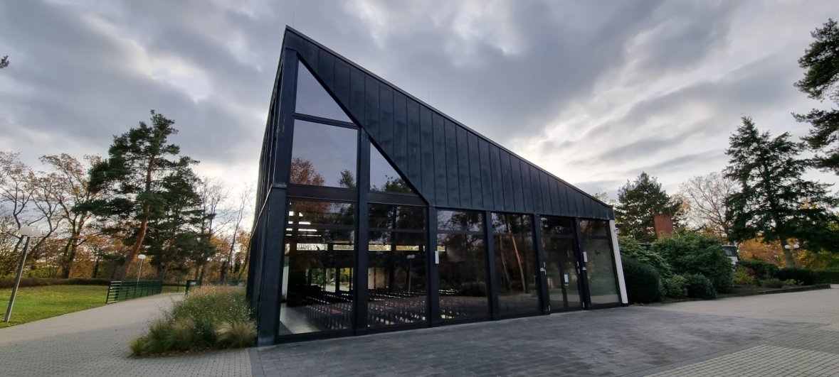 Jügesheim orman mezarlığındaki mezarlık salonu Orman mezarlığının ağaçları ve yürüyüş yolları ile çevrili, siyah çatılı ve büyük cam cepheli modern, konik mezarlık salonu binası.