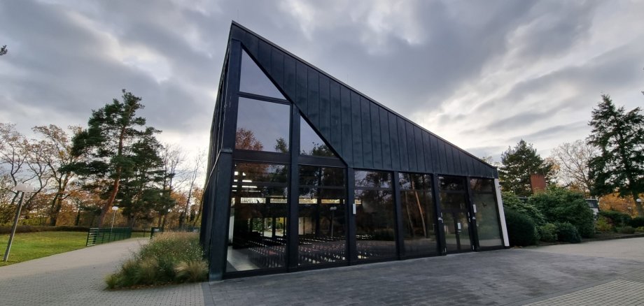 Jügesheim orman mezarlığındaki mezarlık salonu Orman mezarlığının ağaçları ve yürüyüş yolları ile çevrili, siyah çatılı ve büyük cam cepheli modern, konik mezarlık salonu binası.