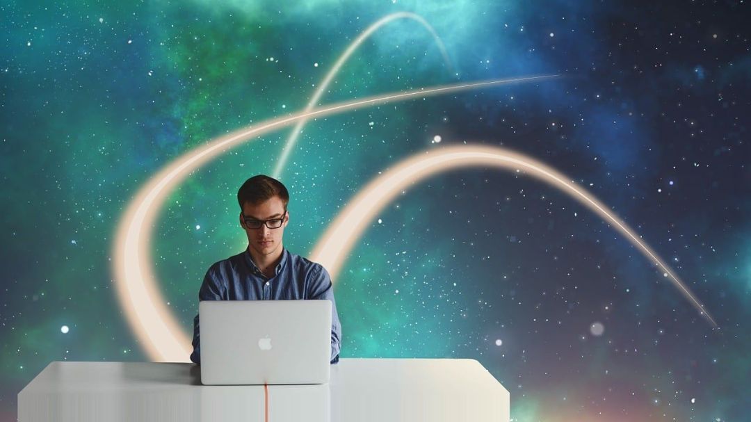Gözlüklü bir adam, yıldızlar ve parlayan çizgilerden oluşan bir arka planın önünde dizüstü bilgisayarının başında oturuyor