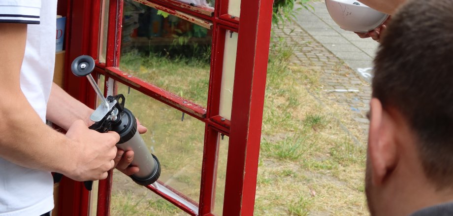 16 Plus Aksiyon Genç bir adam kırmızı ahşap bir kapıyı tutarken, kolu dövmeli bir başka kişi kapıyı sabitliyor ve üçüncü bir kişi de tripod üzerinde akıllı telefon tutuyor