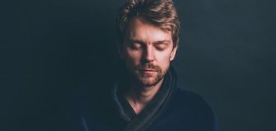 Людина сидить, схрестивши ноги на подушці, руки перед серцевою чакрою в положенні мудри, темний одяг, темний фон.