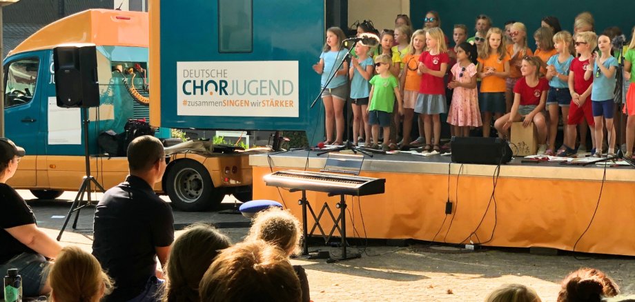 SingBus Дитячий хор співає на сцені перед синьою вантажівкою з написом "DEUTSCHE CHORJUGEND #zusammenSingen wir STÄRKER