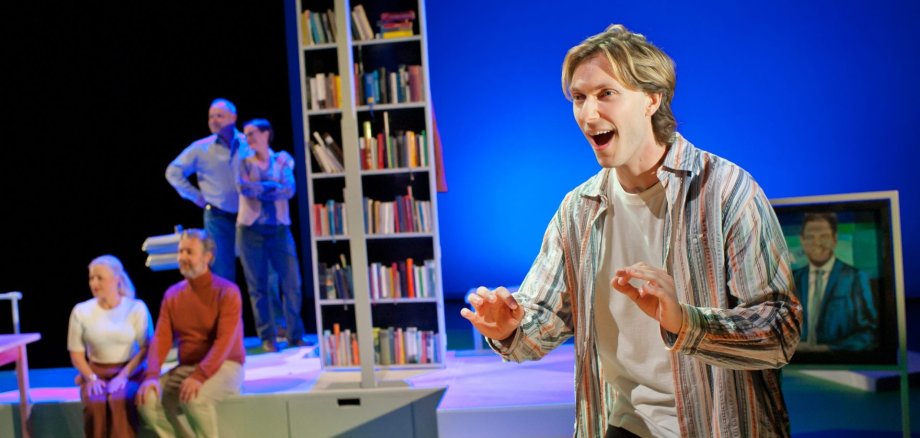 П'єса «Німець» Чоловік у смугастій сорочці жваво жестикулює на сцені перед групою людей, що сидять і стоять, а також перед книжковою шафою та телевізором, на екрані якого зображено чоловіка