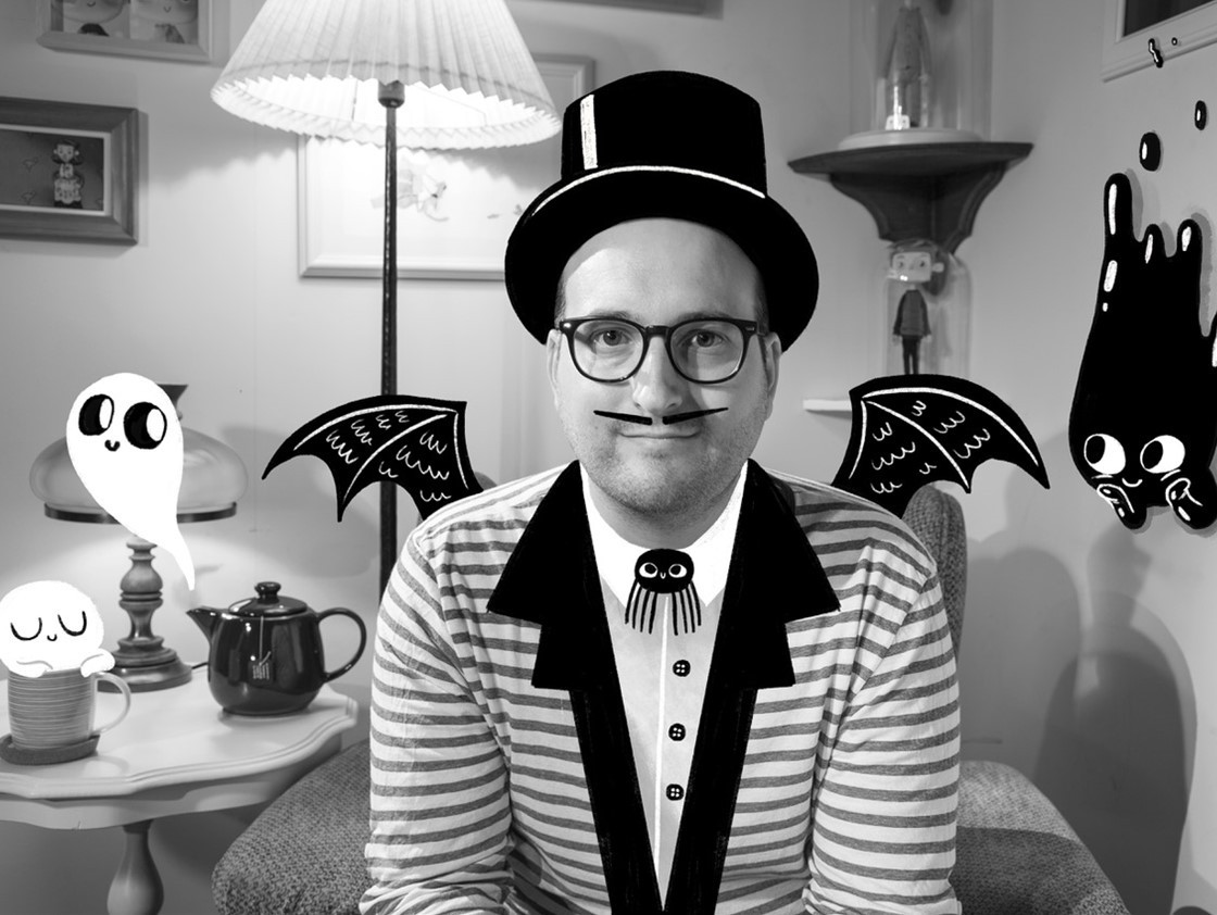 Людина в капелюсі з паперовими крилами, у смугастій сорочці сидить у кімнаті зі світильниками та фігурками на стінах