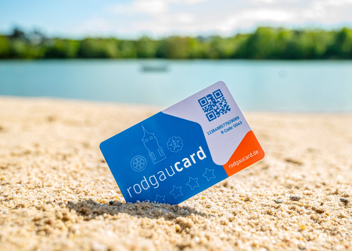 Картка Rodgaucard, встромлена по діагоналі в пісок біля лідо, з розмитою водою і зеленою прибережною рослинністю на задньому плані