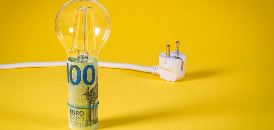 Eine Glühbirne aus Glas steht auf einem 100-Euro-Schein, der zusammengewickelt ist. Neben der Glühbirne liegt ein weißes Stromkabel mit einem Stecker.