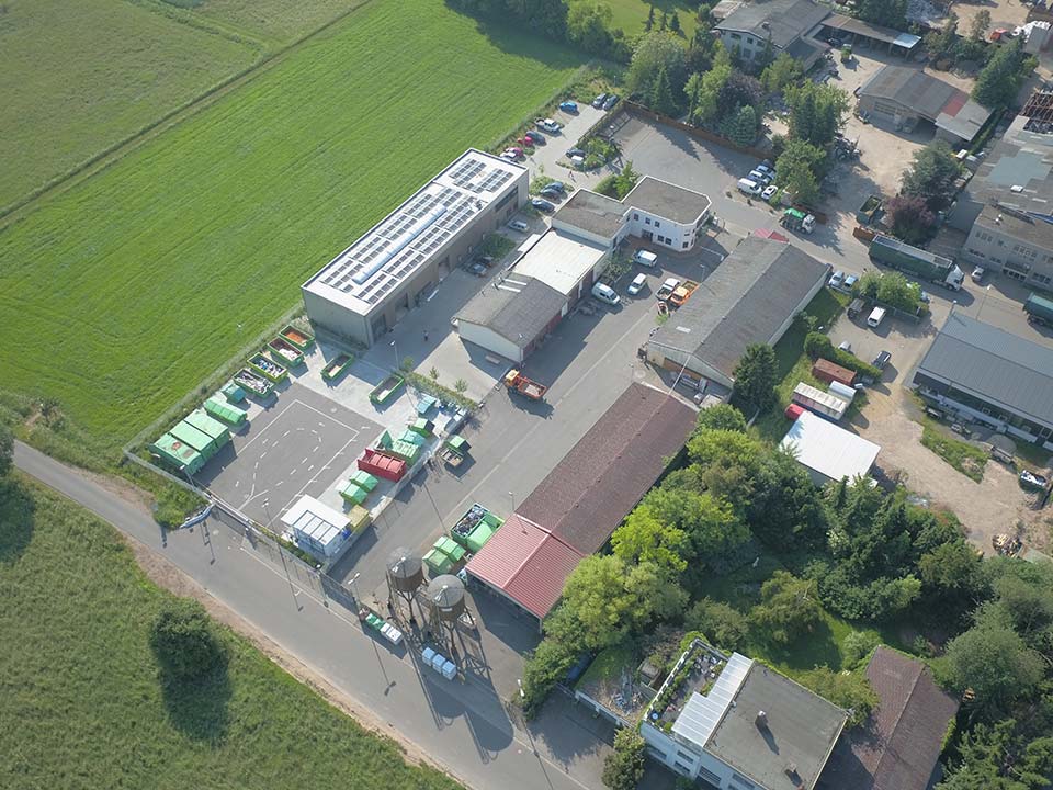 Luftaufnahme eines Wertstoffhofs mit mehreren Gebäuden, Parkplätzen und Abfallcontainern in verschiedenen Farben. Umgeben von Wiesen und Bäumen.