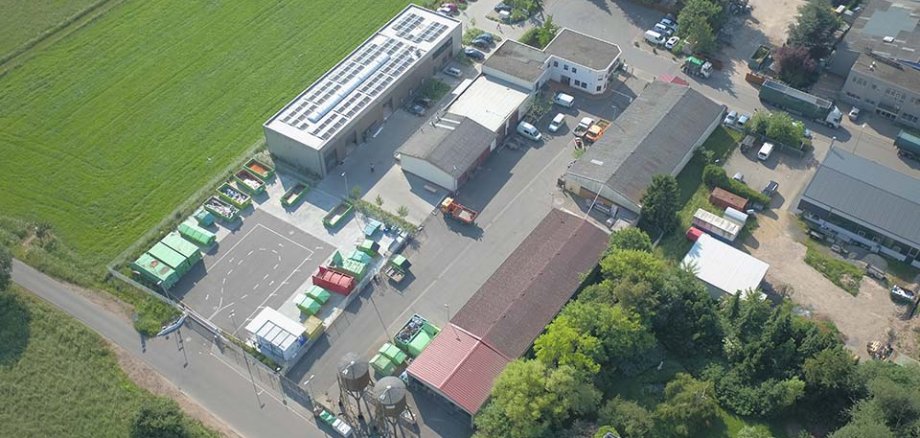 Luftaufnahme eines Wertstoffhofs mit mehreren Gebäuden, Parkplätzen und Abfallcontainern in verschiedenen Farben. Umgeben von Wiesen und Bäumen.