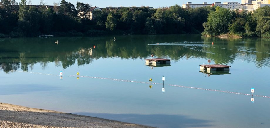 Blick das Strandbad mit zwei schwimmenden Plattformen. Am Ufer Sandstrand und grüne Wiese. Im Hintergrund Bäume und Wohngebäude.