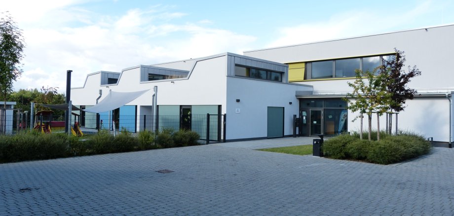 Modernes, weißes Gebäude des Familienzentrums mit gepflastertem Vorplatz, mehreren Fenstern und einem kleinen Baum davor.