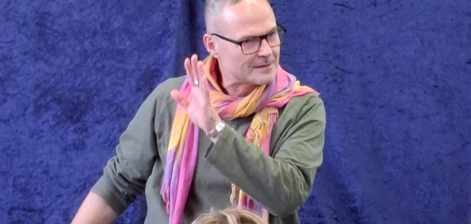 Märchenerzähler Christian Mayer-Glauninger  sitzt vor einer blauen Decke auf dem Boden, trägt einen bunten Schal und gestikuliert mit der rechten Hand vor einer Gruppe von Kindern.