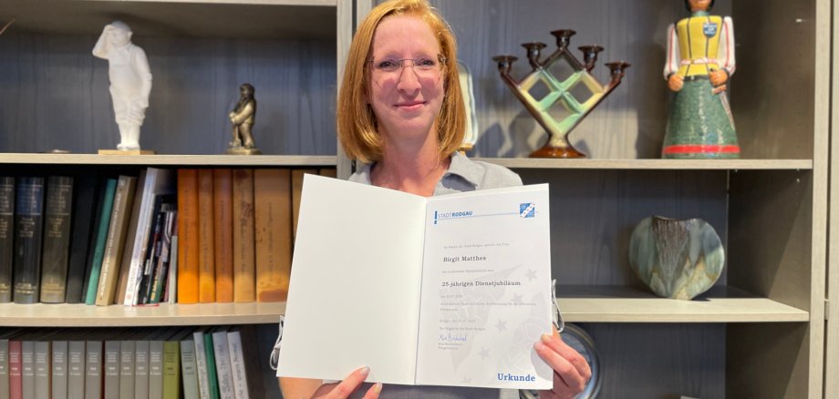 Birgit Mattes hält eine Urkunde mit dem Text '25 Jahre Dienstjubiläum'. Im Hintergrund stehen Bücher, eine Statue und eine dekorative Glasvitrine.