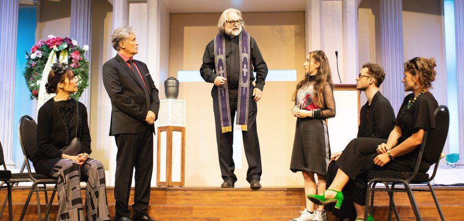 Bühnenbild aus dem Theaterstück "Kalter weißer Mann" mit sechs Personen, eine steht auf einer kleinen Bühne in schwarzer Kleidung und violettem Priesterschal, die anderen sitzen oder stehen davor, Säulen, Urne  und Blumenkranz im Hintergrund.