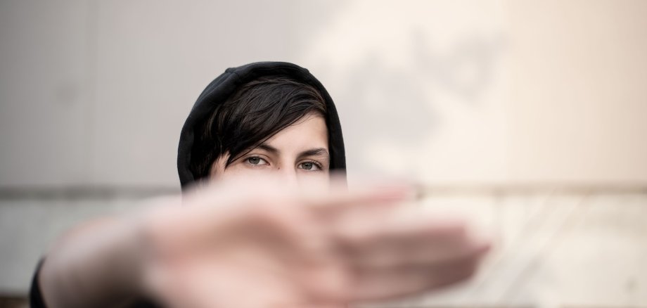 Frau mit schwarzer Kapuzenjacke hält Handfläche zur Kamera, unscharfer Hintergrund mit Wand und Graffiti