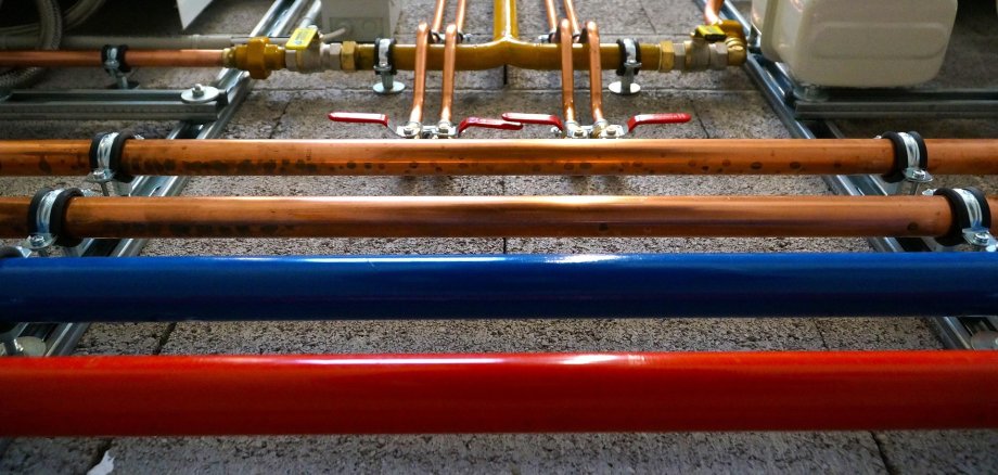 Rohrleitungen in verschiedenen Farben (rot, blau, orange) mit Ventilen und Verbindungsstücken auf einer Betonwand installiert.