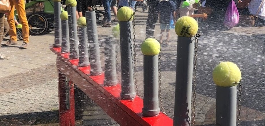 Eine rote Wasserstation mit mehreren grauen Rohren, die mit Tennisbällen an Ketten versehen sind. Im Hintergrund sind viele Menschen auf einem Fest.