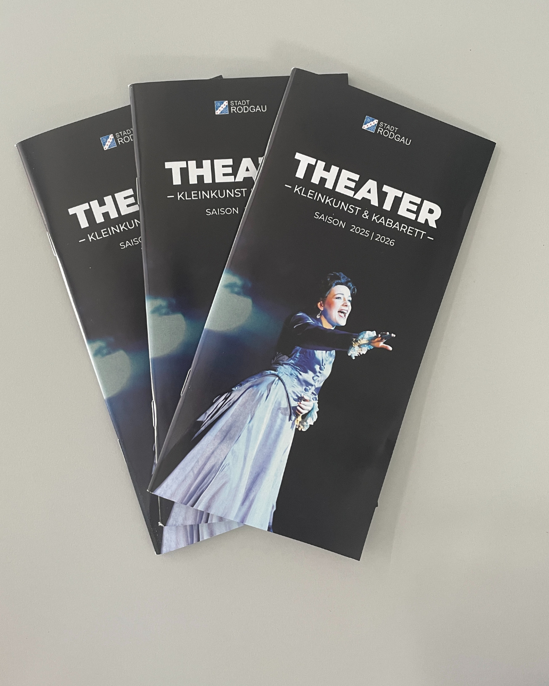 Kulturhighlights 2025/26 Drei schwarze Theaterbroschüren mit dem Titel 'THEATER - KLEINKUNST & KABARET' für die Saison 2025/2026. Eine Person in einem weißen Kostüm ist abgebildet.