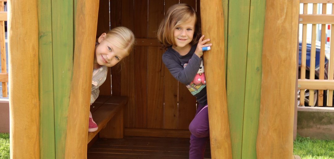 Holzspielhaus mit grün gestrichenen Wänden und einer offenen Tür. Zwei Kinder stehen im Eingang, eines schaut heraus. Eine kleine Treppe führt zur Tür.