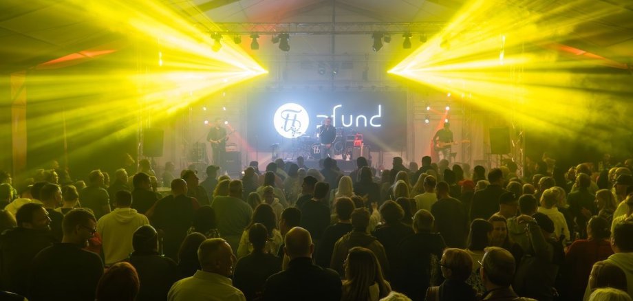 Eine Menschenmenge steht in einem Zelt und schaut auf eine Band, die auf einer Bühne spielt. Gelbe Lichtstrahlen strahlen von der Decke auf die Zuschauer.