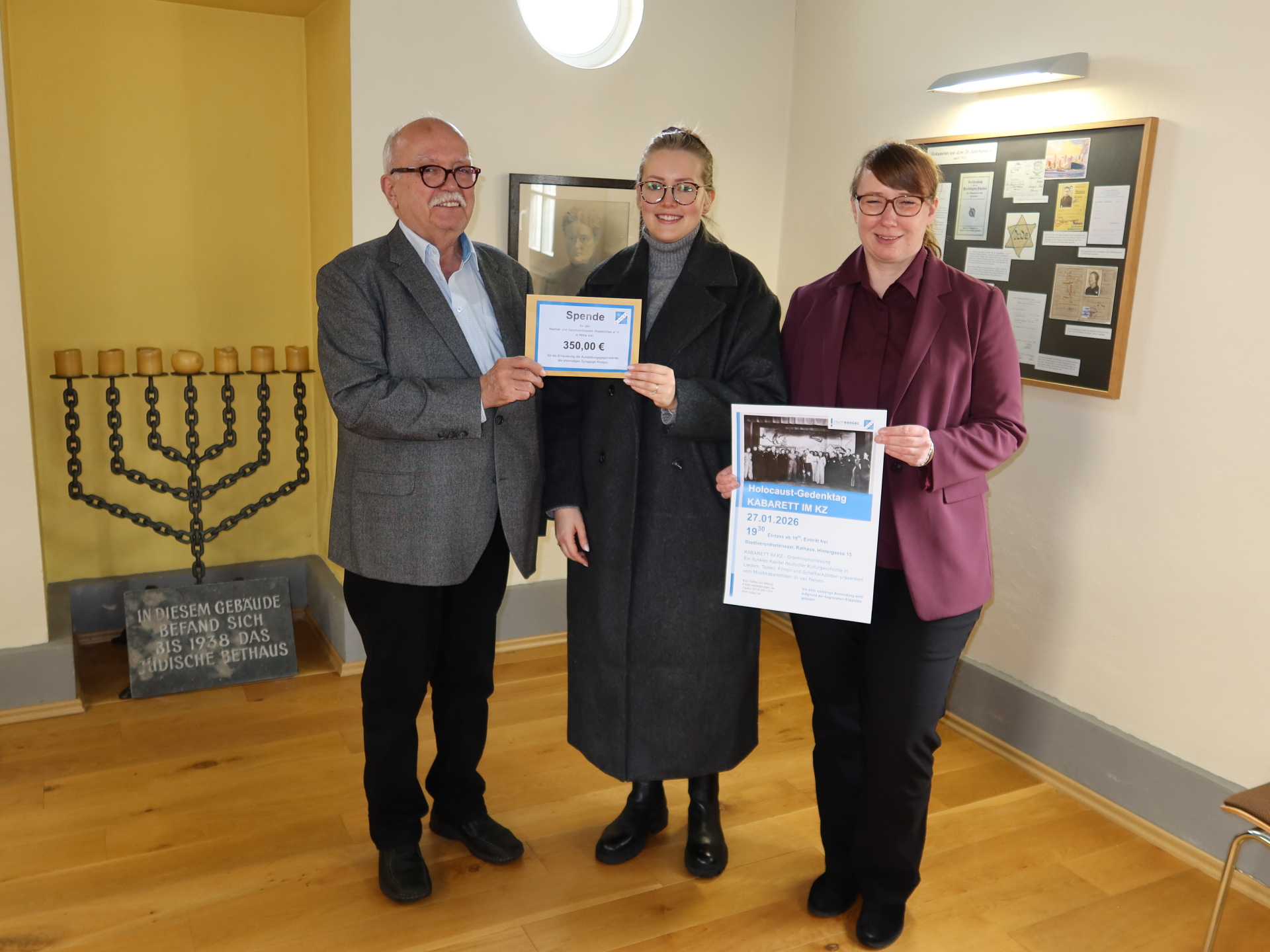 Drei Personen stehen in einem Raum mit einem Menora-Leuchter und einer Gedenktafel und halten ein Schild mit der Aufschrift 'Spende € 350,00' sowie ein Plakat zur Holocaust-Gedenkveranstaltung.