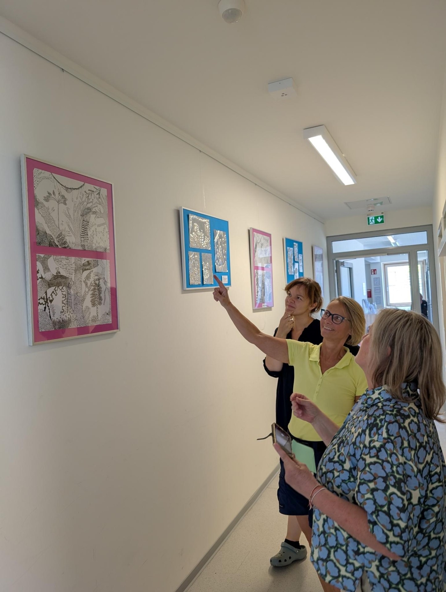Bilder der Geschwister-Scholl-Schule im Familienzentrum Drei Frauen betrachten und zeigen auf gerahmte Bilder an einer weißen Flurwand in einem Familienzentrum