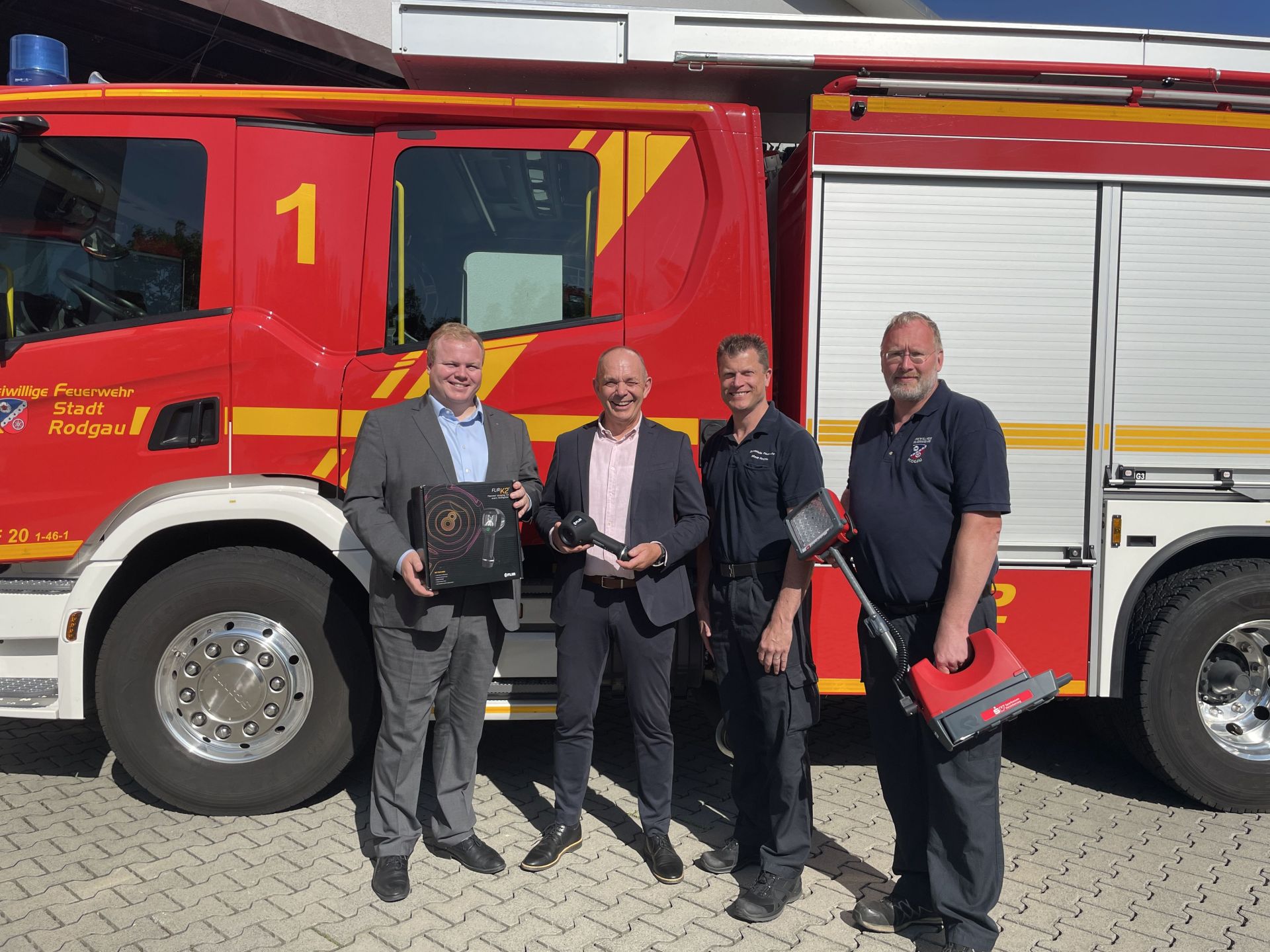 Vier Männer stehen vor einem roten Feuerwehrfahrzeug der Freiwilligen Feuerwehr Stadt Rodgau, zwei halten Wärmebildkameras, zwei tragen Anzüge, zwei tragen Feuerwehruniformen.
