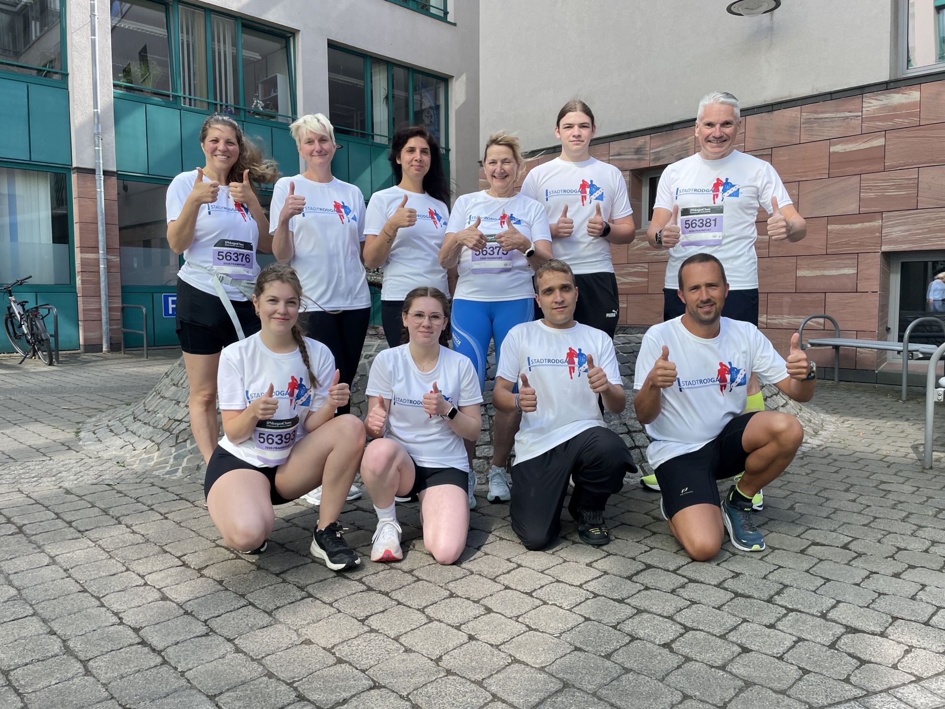 Gruppe von neun Mitarbeitenden der Stadt in weißen T-Shirts mit JP Morgan Chase-Logo posiert mit Daumen hoch im gepflastertem Rathausinnenhof
