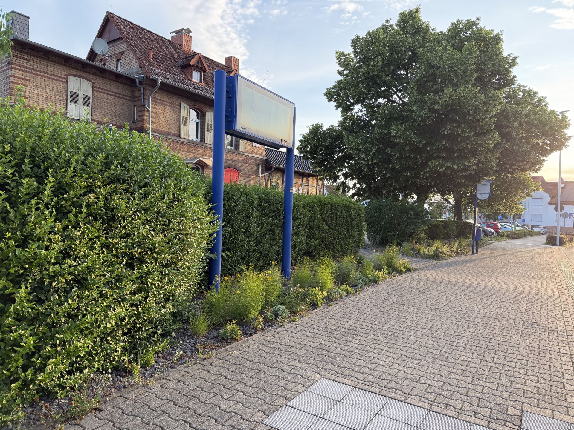 Blick auf den Bahnhof Nieder-Roden Blick auf den Bahnhof Nieder-Roden mit einem blauen Schild vor einer Hecke, einem gepflasterten, sauberen Gehweg und einem großen Baum im Hintergrund
