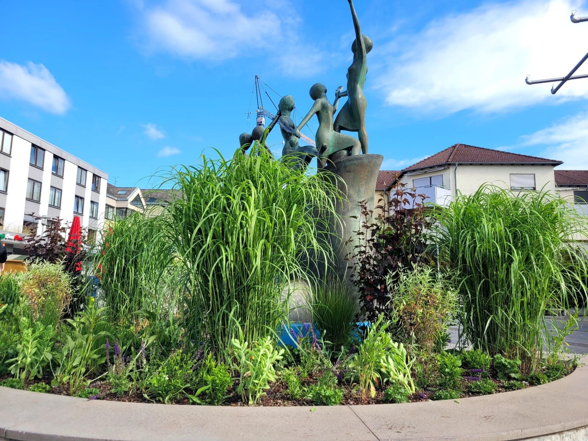 Begrünter Brunnen auf dem Puiseauxplatz Brunnen auf dem Puiseauxplatz mit einer Skulptur aus mehreren Figuren, umgeben von hohen Gräsern und Pflanzen, im Hintergrund Wohn- und Geschäftshäuser