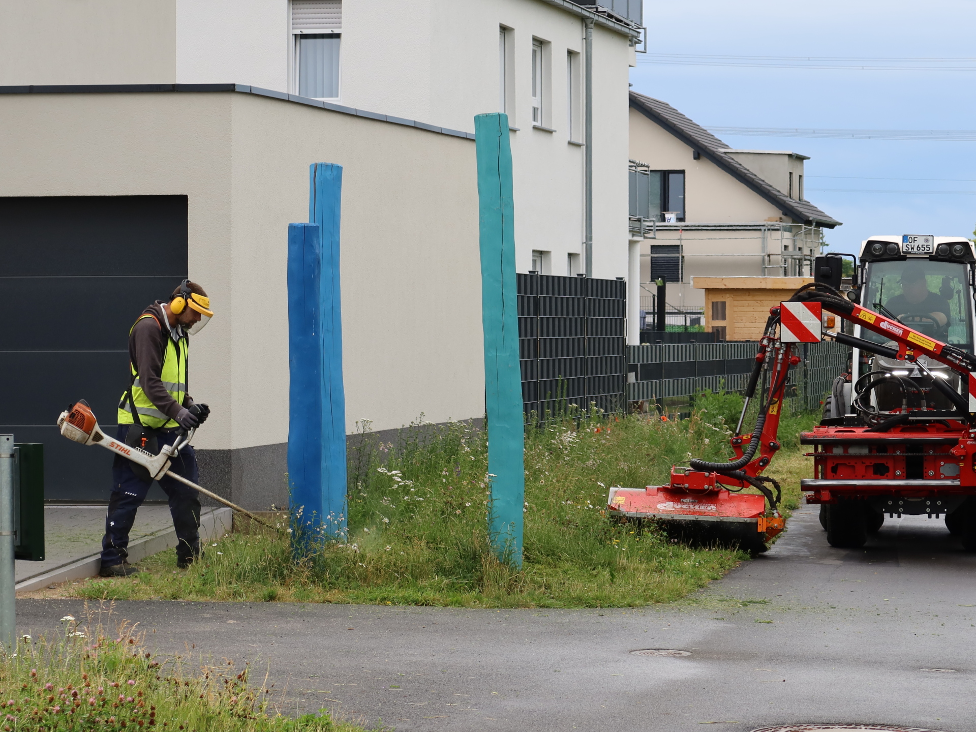 Grünpflege Stadtwerke Rodgau Mitarbeiter der Grünpflege Stadtwerke Rodgau mäht mit Motorsense an einem Wohnhaus neben zwei blauen Holzpfosten, rechts steht ein Mähgerät an einem Traktor auf einem Fuß-/Radweg.