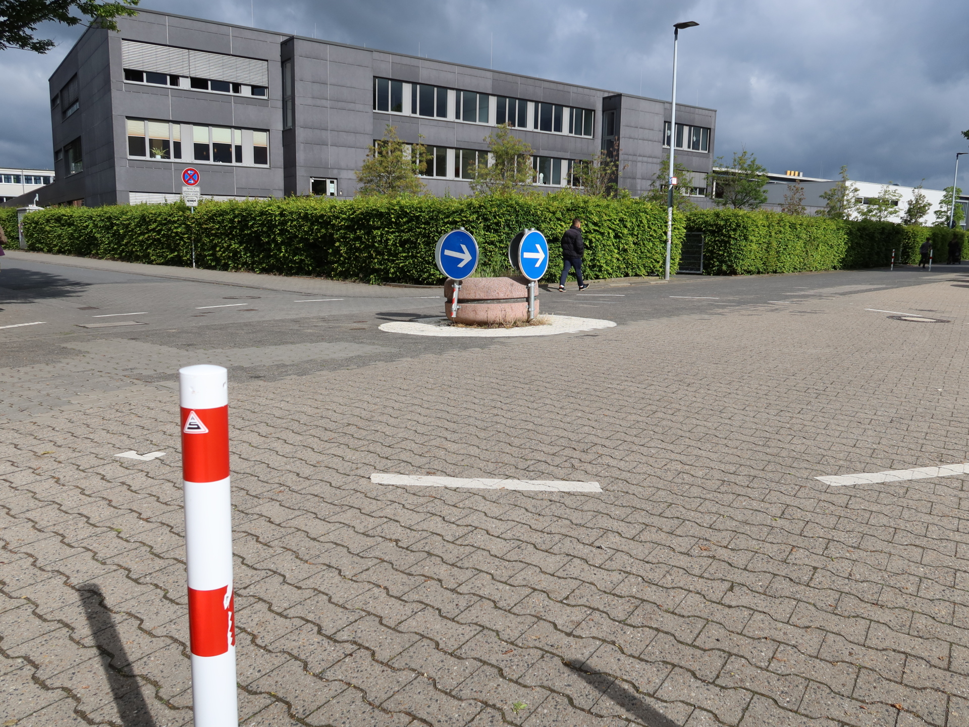 Kleiner Kreisverkehr zwischen Kita 18 und Georg-Büchner-Schule Kleiner Kreisverkehr mit zwei blauen Richtungspfeilen auf einem runden Mittelinselpodest zwischen Kita 18 und Georg-Büchner-Schule, umgeben von gepflastertem Boden und dem mehrstöckigen Gebäude der Schule im Hintergrund