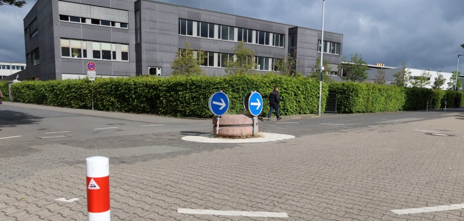 Kleiner Kreisverkehr zwischen Kita 18 und Georg-Büchner-Schule Kleiner Kreisverkehr mit zwei blauen Richtungspfeilen auf einem runden Mittelinselpodest zwischen Kita 18 und Georg-Büchner-Schule, umgeben von gepflastertem Boden und dem mehrstöckigen Gebäude der Schule im Hintergrund