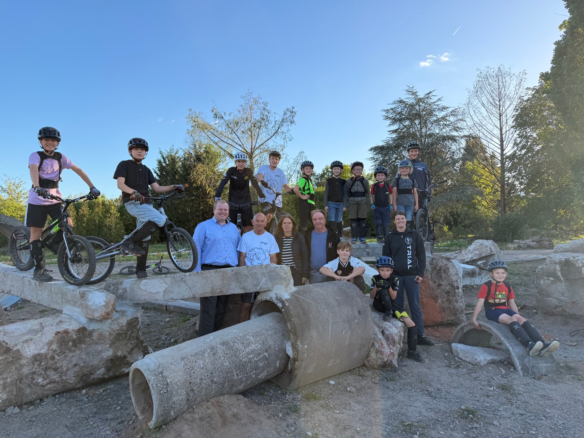 Neues Gelände für die TSV Trail-Abteilung Gruppe von Kindern und Erwachsenen mit Helmen und Fahrrädern auf und um große Betonrohre und Felsen im Freien