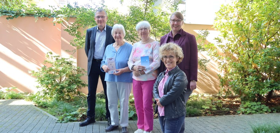 Gewinnerinnen des Seniorentag-Gewinnspiels Fünf Personen stehen in einem Garten auf gepflastertem Weg, drei ältere Frauen halten kleine Broschüren, zwei jüngere Personen stehen seitlich und hinten