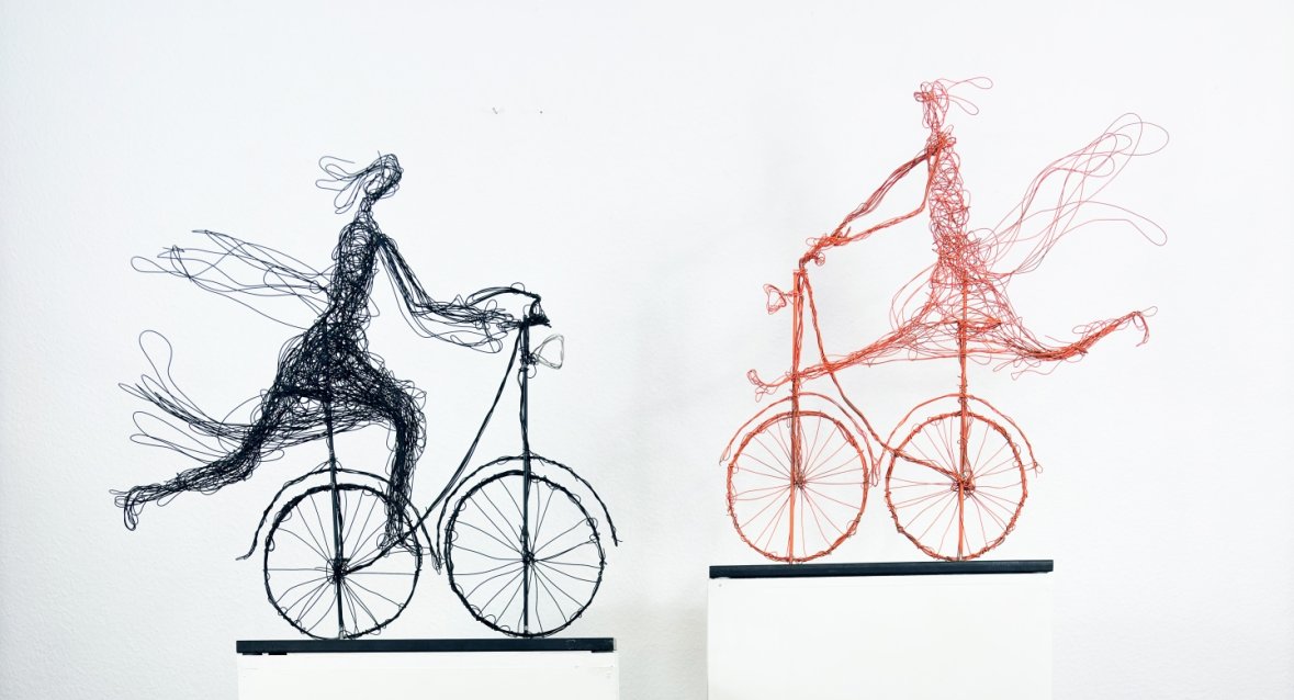 Kunstwerk von Henriette Tomasi Zwei filigrane Drahtskulpturen von Figuren auf Fahrrädern auf weißen Podesten vor weißer Wand