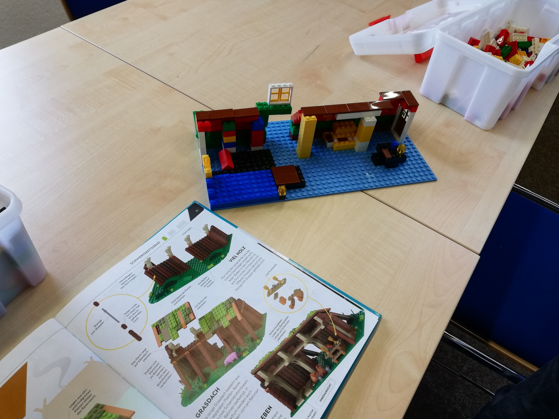 Große Kinder-Bauaktion in der Stadtbücherei Nieder-Roden Auf einem Tisch steht ein teilweise gebautes Lego-Modell auf einer blauen Grundplatte neben einer geöffneten Bauanleitung und zwei weißen Behältern mit Lego-Steinen.