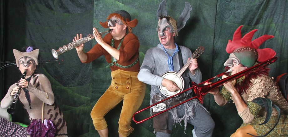 Theatermärchen "Die Bremer Stadtmusikanten" Vier Personen in fantasievollen Tierkostümen spielen verschiedene Blasinstrumente und eine Banjo-ähnliche Gitarre auf einer Bühne mit grünem Hintergrund