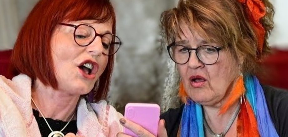 "Gerda & Lisbeth" auf dem Frauentag Zwei Frauen mit Brille schauen gemeinsam auf ein Smartphone, die eine zeigt etwas darauf und spricht, die andere hört zu