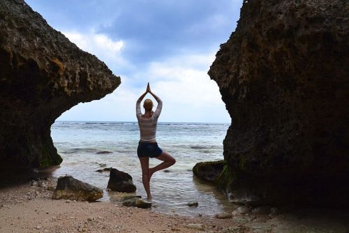 vhs Yoga für Nacken, Schultern und den Rücken Person steht im Baum-Yoga auf einem Felsen am Strand zwischen zwei großen Felsen mit Blick auf das Meer