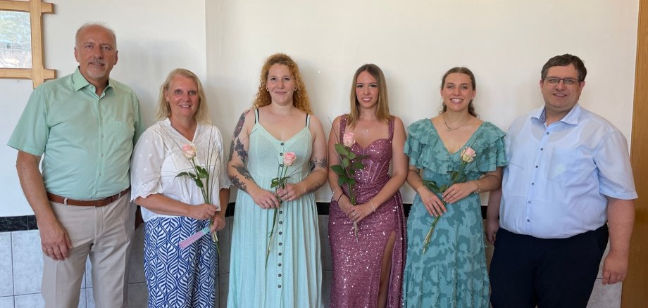 Drei städtische Auszubildende nach bestandener Prüfung zur Verwaltungsfachangestellten mit Ausbildern Sechs Personen stehen nebeneinander in einem Raum mit weißer Wand und einem Kreuz an der linken Wand, drei Frauen in Kleidern halten jeweils eine Rose, zwei Männer tragen Hemden und Hosen