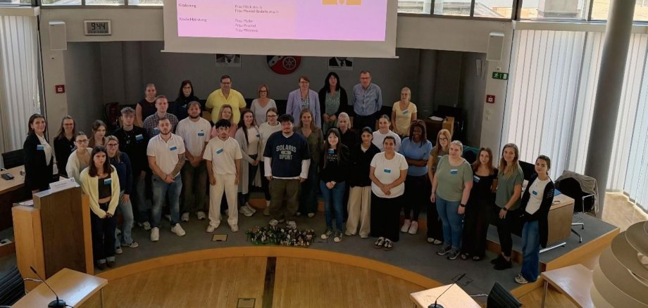 Neue pädagogische Auszubildende 2026 Gruppe von Erwachsenen steht im Stadtverordnetensitzungssaal mit großen Fenstern und einer Präsentation an der Wand