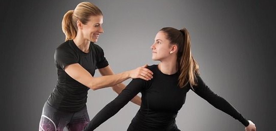 Rückenfit Zwei Frauen in Sportkleidung, eine steht in einer tiefen Kniebeuge mit ausgestreckten Armen, die andere berührt unterstützend ihre Schulter