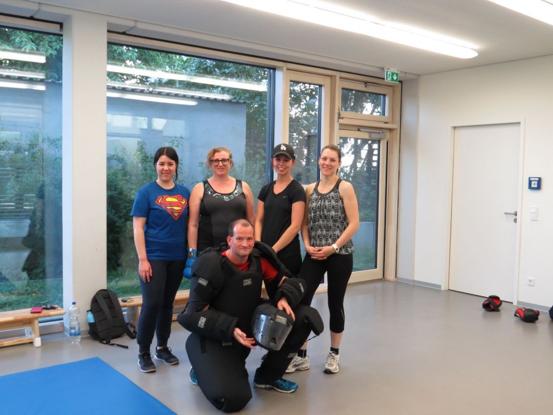 Vier Frauen in Sportkleidung stehen in einem hellen Raum hinter einem Mann in schwarzer Schutzkleidung, der einen Helm hält