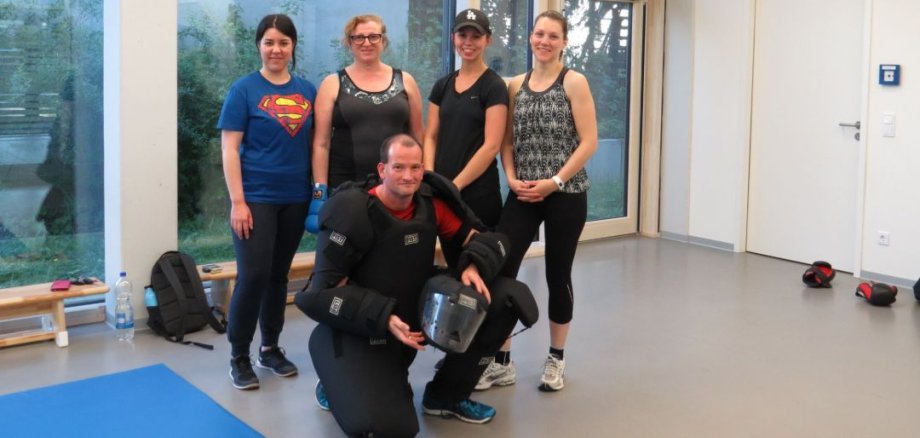 Vier Frauen in Sportkleidung stehen in einem hellen Raum hinter einem Mann in schwarzer Schutzkleidung, der einen Helm hält